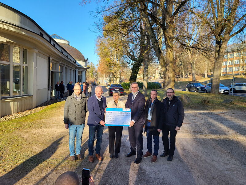 Gruppenfoto unter Bäumen, eine Frau in der Mitte, umgeben von 5 Männern, hält symbolischen Scheck in der Hand