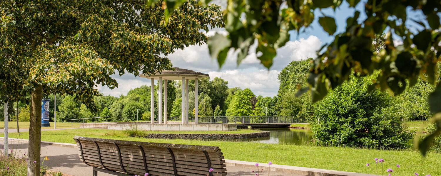 große Sitzbank unter Bäumen im Park, im Hintergrund See und ein stilisierter Brunnentempel
