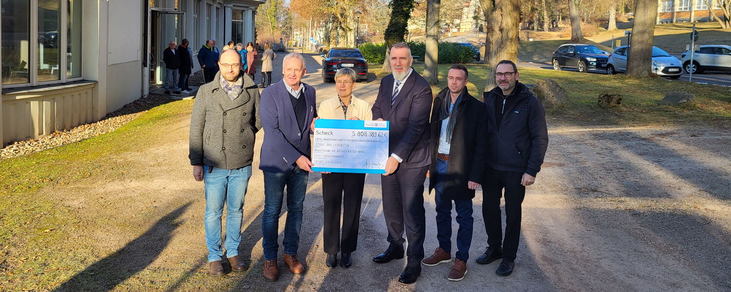 Gruppenfoto unter Bäumen, eine Frau in der Mitte, umgeben von 5 Männern, hält symbolischen Scheck in der Hand