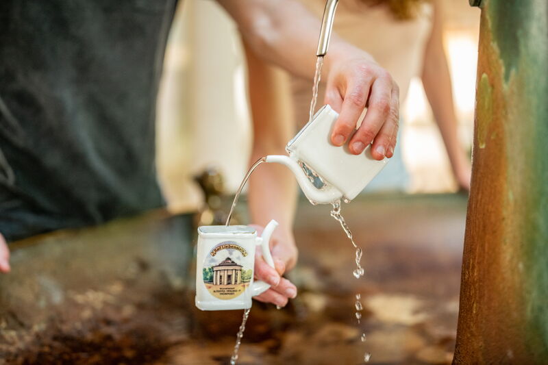 Heilwasser fließt in zwei Kurbecher, gehalten von jeweils einer Hand
