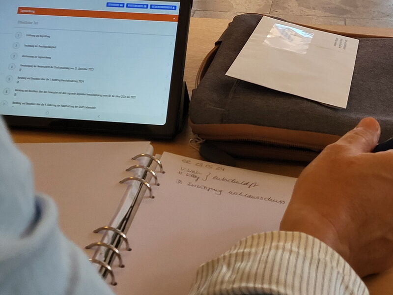 Tisch mit Tablet, auf dem eine Tagesordnung angedeutet ist, Notizblock und Person, die Stift in der Hand hält