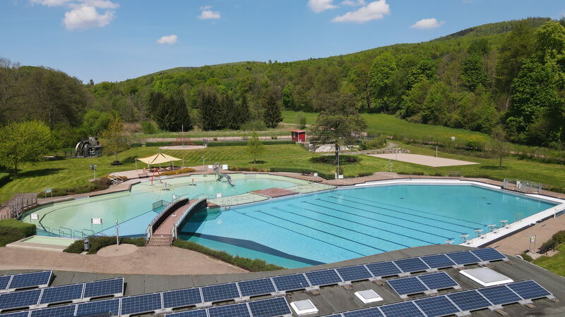 Luftaufnahme des Freibades mit Schwimmbecken, Nichtschwimmerbecken, Liegewiesen und Bergen im Hintergrund