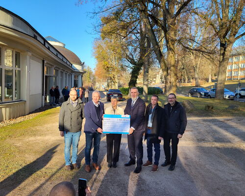 Gruppenfoto unter Bäumen, eine Frau in der Mitte, umgeben von 5 Männern, hält symbolischen Scheck in der Hand