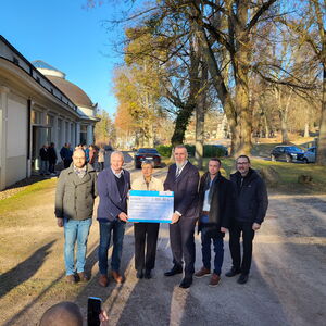 Gruppenfoto unter Bäumen, eine Frau in der Mitte, umgeben von 5 Männern, hält symbolischen Scheck in der Hand