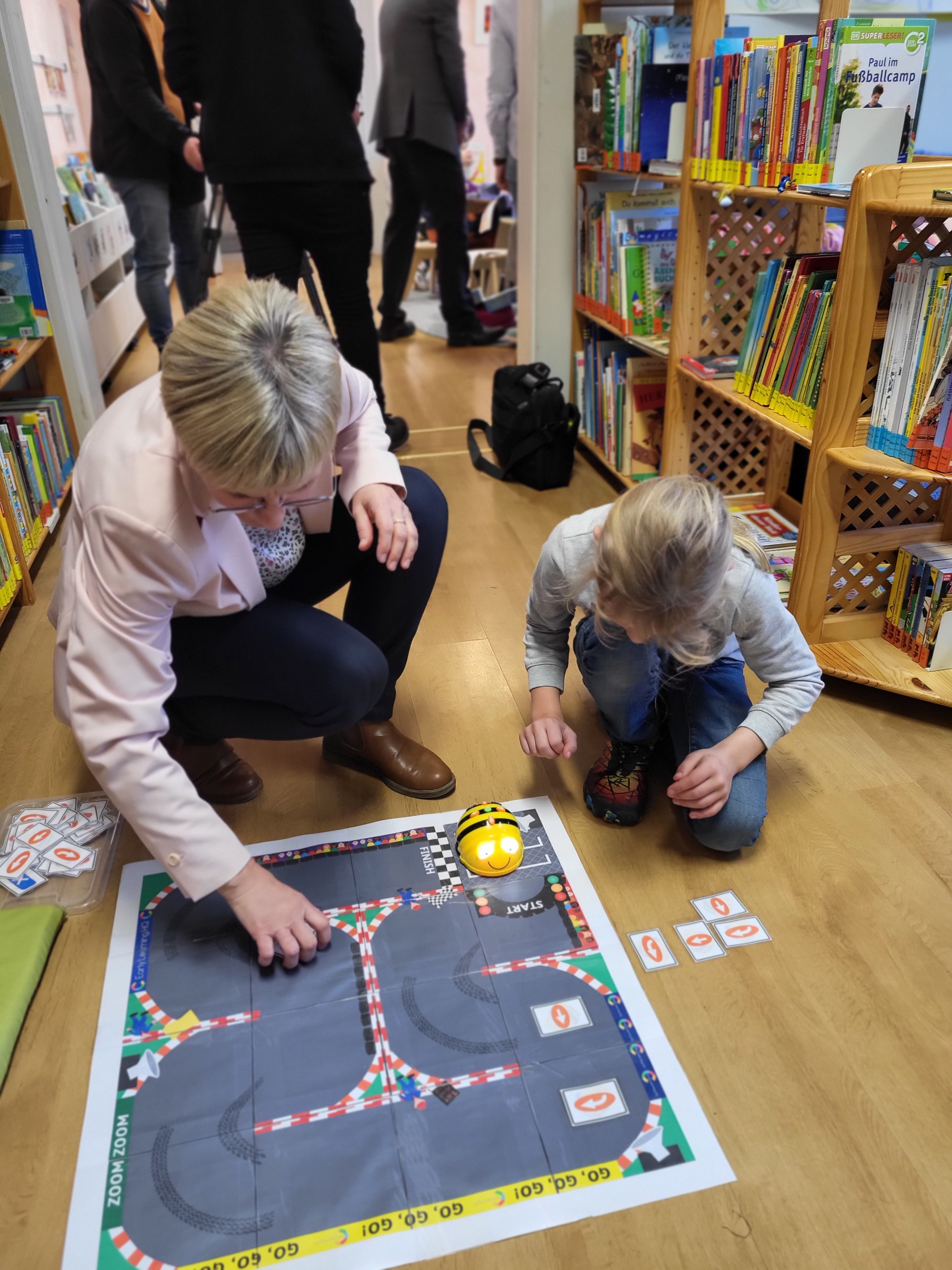 Bee-Bot-Parcour. Frau und Kind knien davor und legen Hilfekärtchen mit Richtungspfeilen.