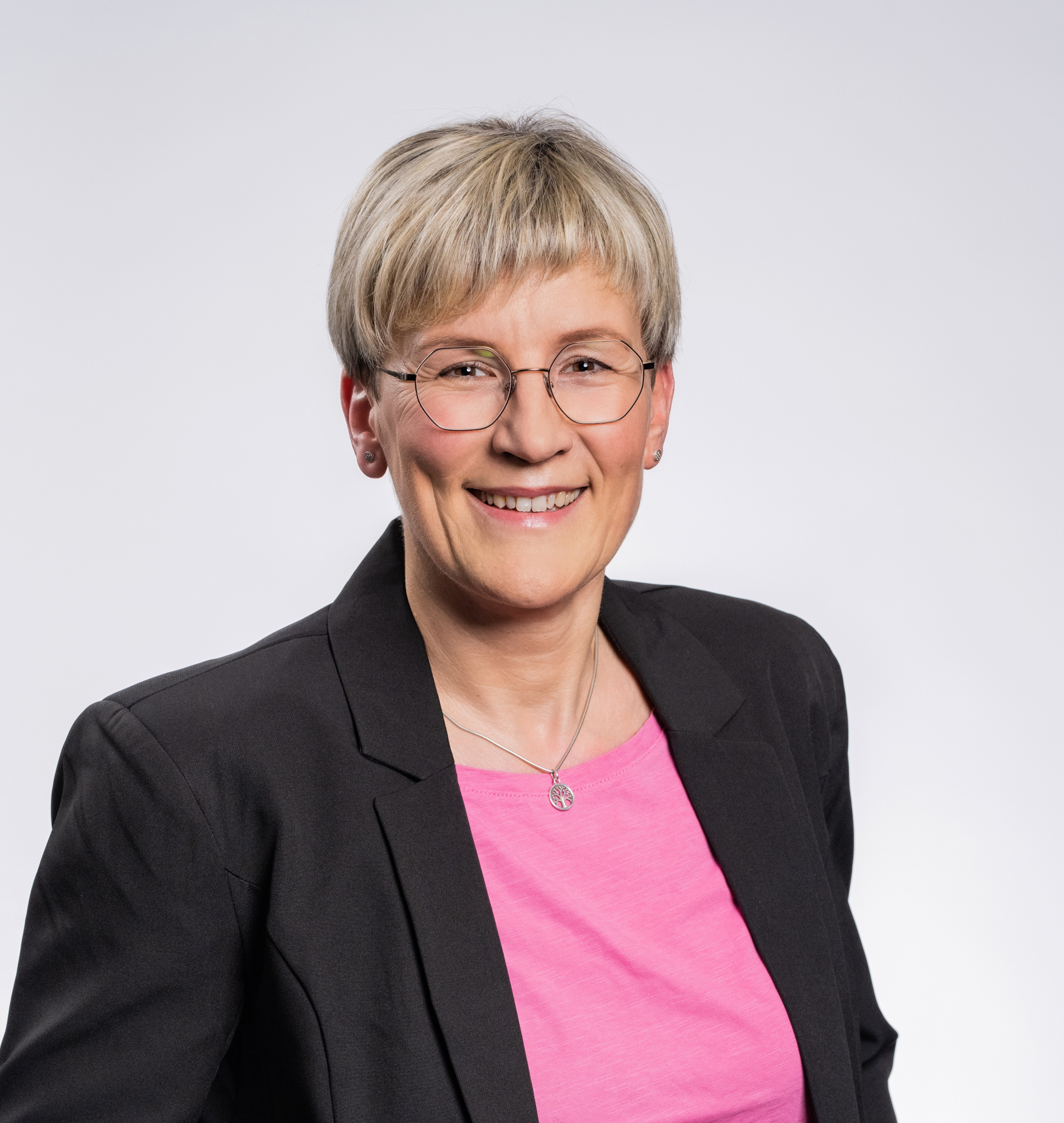 Porträt von Susanne Rakowski: Frau mit blonden kurzen Haaren, Brille, schwarzen Blazer und pinkfarbenen Shirt